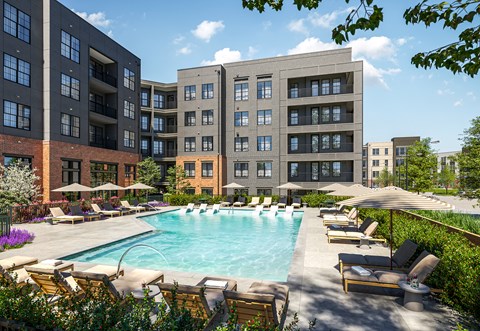 A large swimming pool surrounded by lounge chairs and umbrellas in front of apartment buildings.
