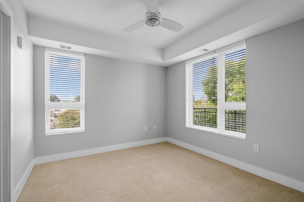 A room with a fan, two windows, and a carpeted floor.