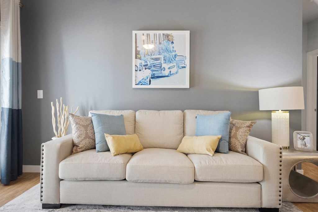 A white couch with blue and yellow pillows in front of a grey wall with a framed picture above it.