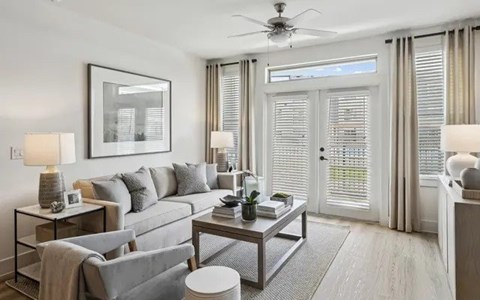 A living room with a grey sofa, a grey armchair, a glass table with a plant on it, and a ceiling fan.