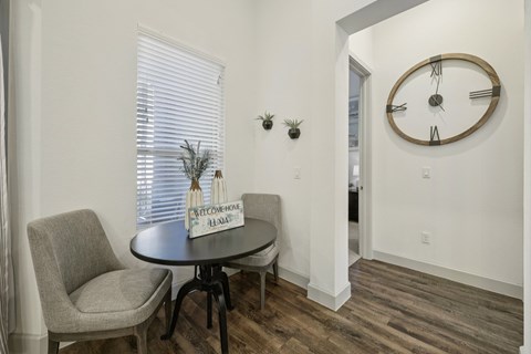A bright front den area located directly off the main entrance, featuring soft eggshell walls and dark wood floors that lead into the rest of the apartment.