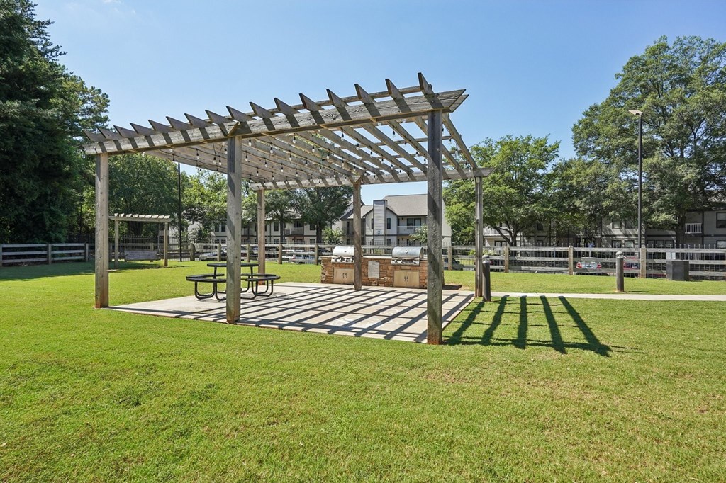 A wooden pergola with a picnic table underneath it is situated in a grassy area.