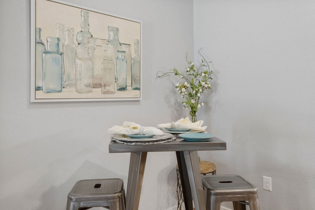 A painting of bottles hangs on a wall above a table with a napkin and a vase of flowers.