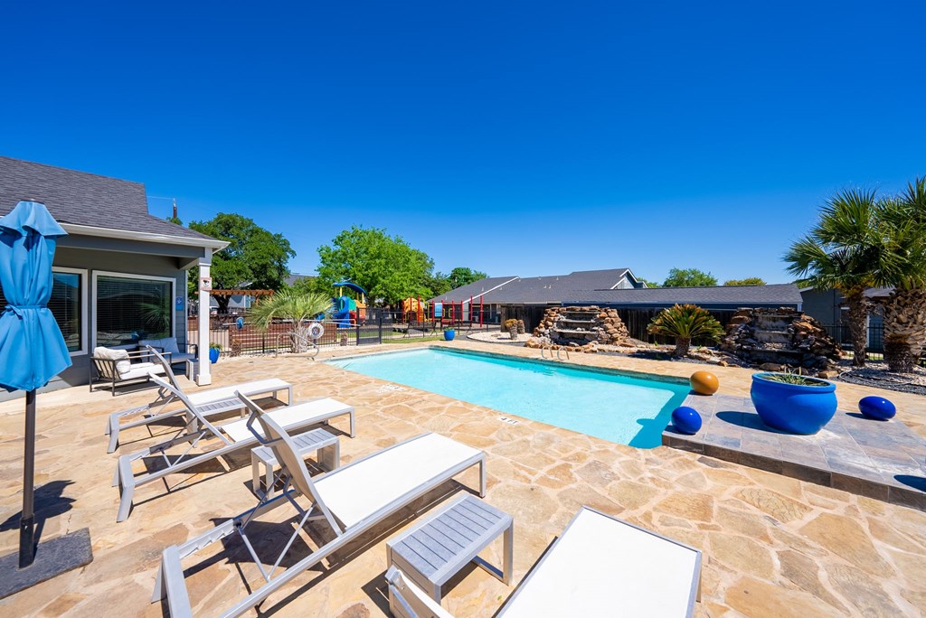 A pool with lounge chairs and umbrellas is surrounded by a stone patio.
