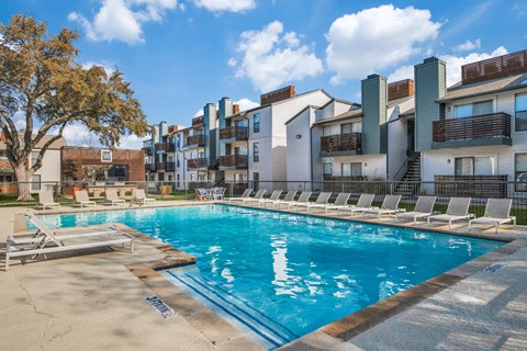 A swimming pool surrounded by lounge chairs and buildings in the background.