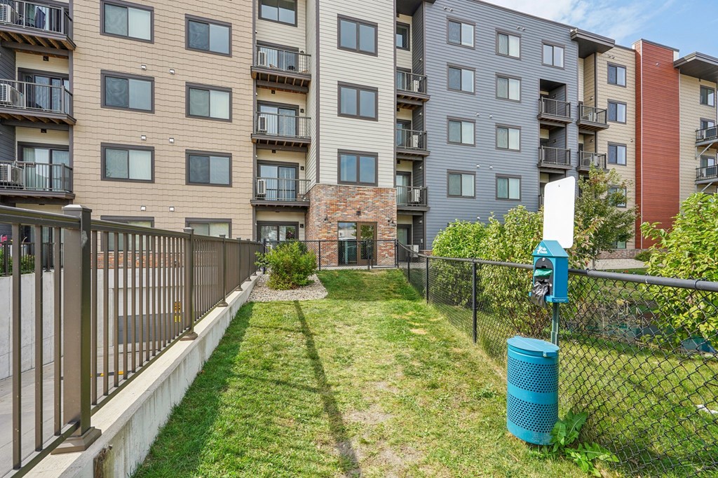 A blue trash can sits in the grass next to a fence in front of apartment buildings.