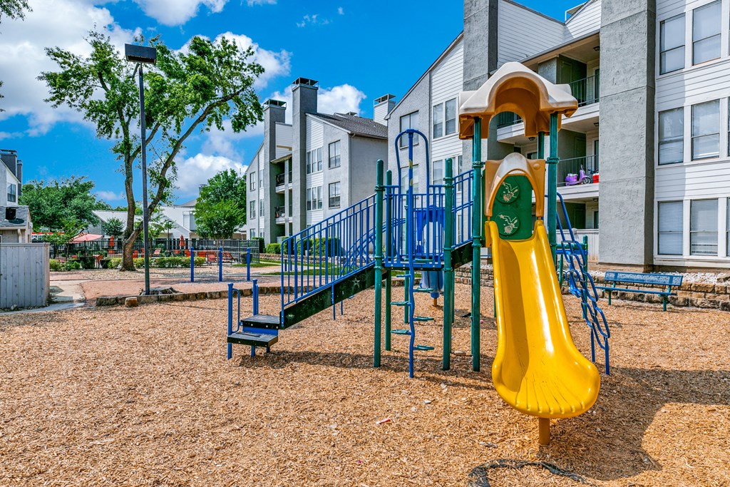 a playground with a yellow slide and blue playset in front of apartments