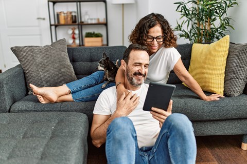 A man and woman are sitting on a grey couch with a tablet in the man's hands.