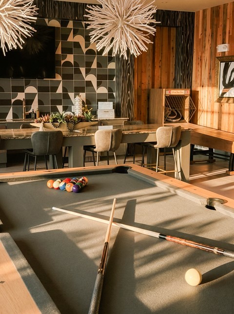 A pool table with a white cue stick and balls is in the foreground of a room with a dining table and chairs.