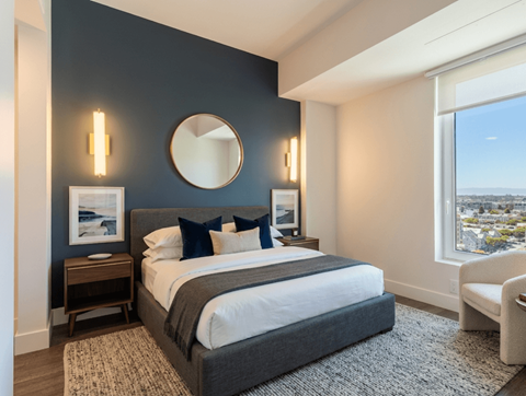 A bedroom with a large bed, a round mirror, and a window overlooking a cityscape.