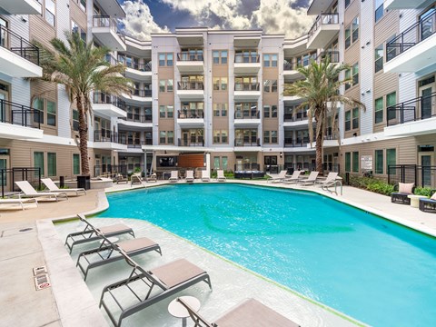 A large swimming pool surrounded by lounge chairs and palm trees in front of a multi-story apartment building.