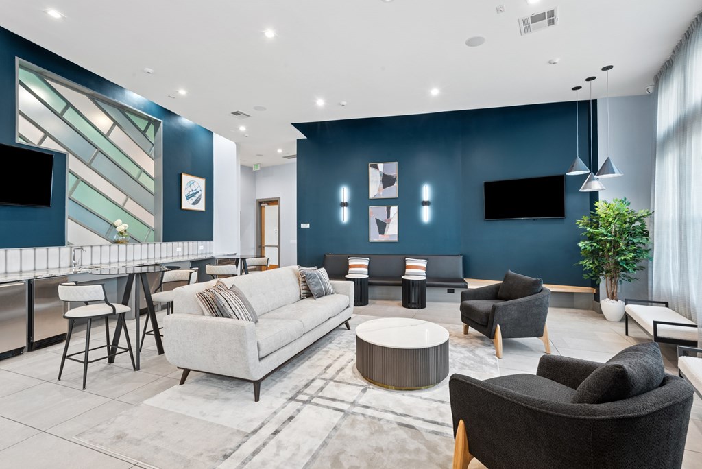 A modern living room with a grey sofa, a black armchair, a grey coffee table, a grey rug, a grey dining table with four chairs, a TV, a plant, and a wall with a geometric design.