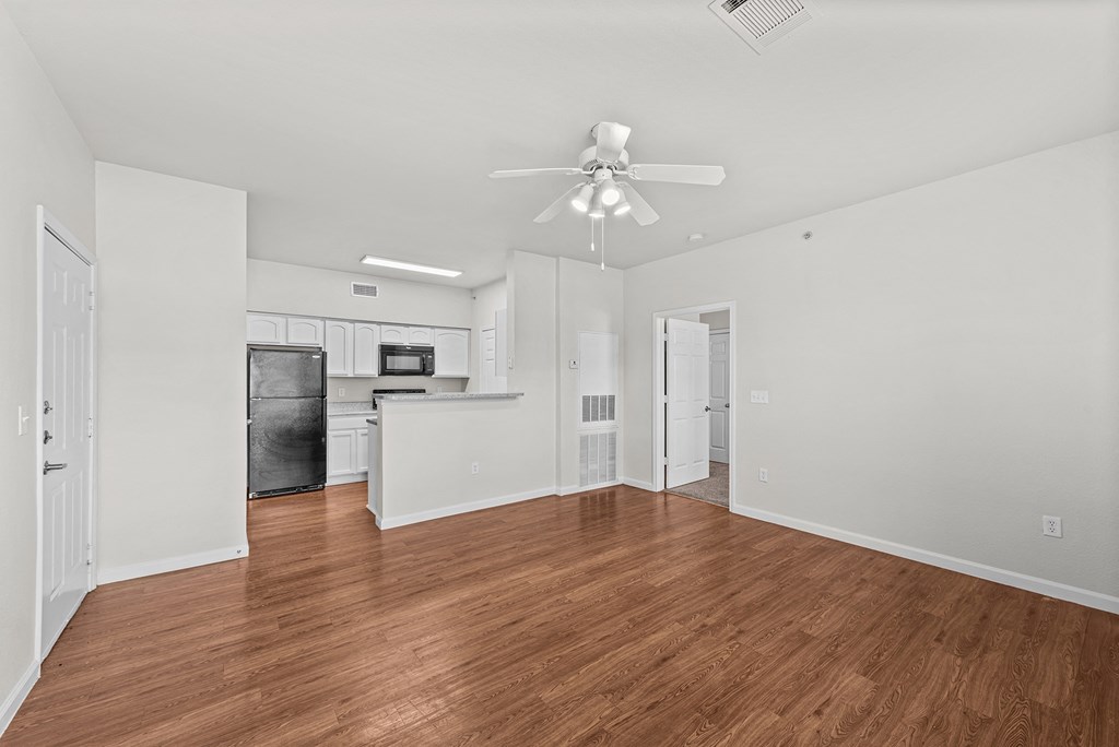 the living room and kitchen of an apartment with wood flooring and a ceiling fan