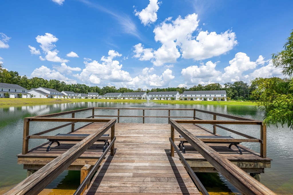 A wooden dock extends into a calm body of water with a row of houses in the distance.