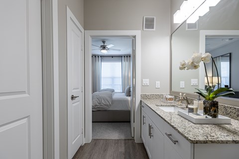 A bathroom with a vanity, mirror, and a bedroom in the background.