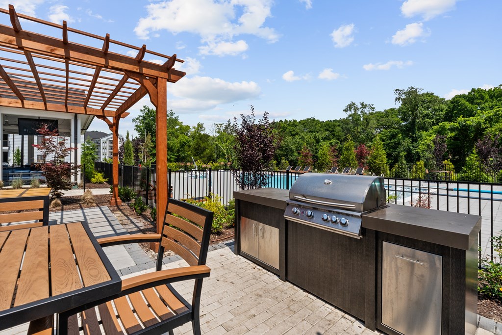 A wooden table and bench are on a patio with a grill and a pool in the background.
