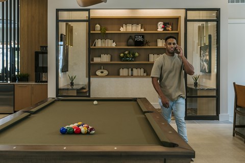 A man is standing by a pool table and talking on a cell phone.