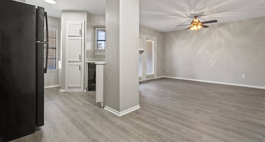 an empty living room with a ceiling fan and a kitchen in the background