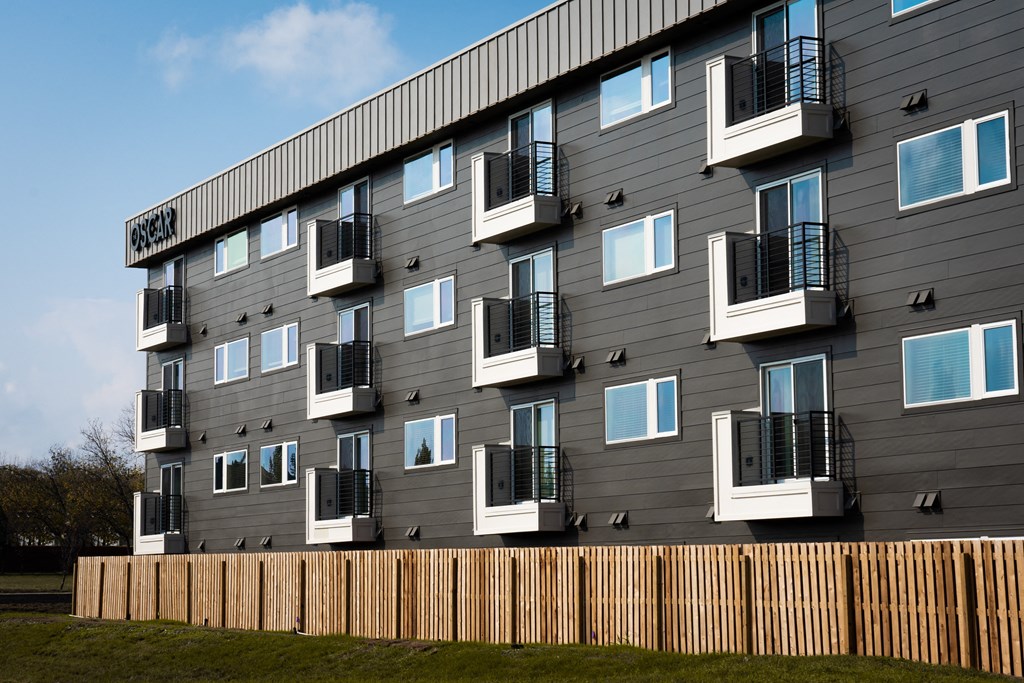 a picture of a grey apartment building with white and black balconies and a wooden fence