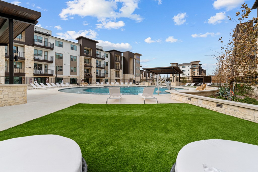 A swimming pool is surrounded by a grassy area and chairs in front of apartment buildings.