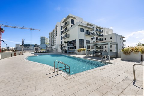 A large swimming pool is surrounded by a tiled patio and a building with balconies.