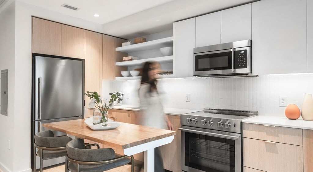 A woman is walking through a modern kitchen with a refrigerator, oven, and microwave.