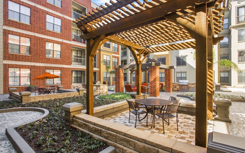 A wooden pergola is situated over a table and chairs in a courtyard.