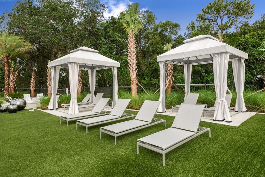 A white gazebo is surrounded by white lounge chairs on a green lawn.