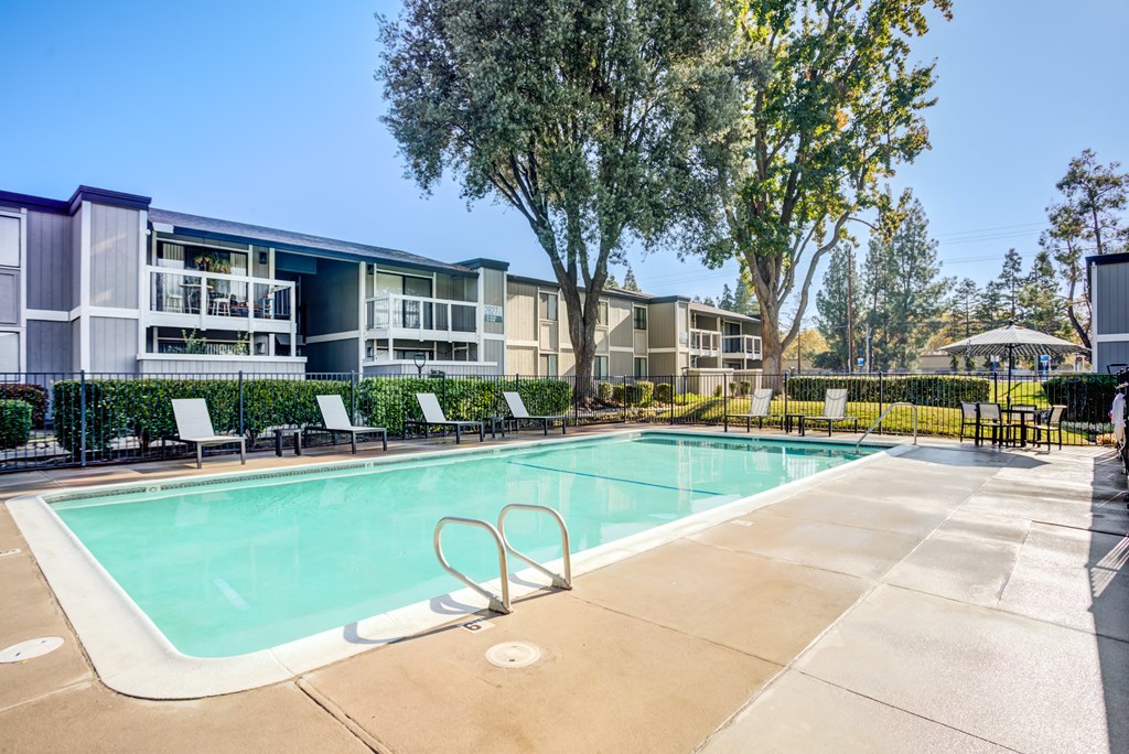 A swimming pool surrounded by trees and chairs in front of apartment buildings.
