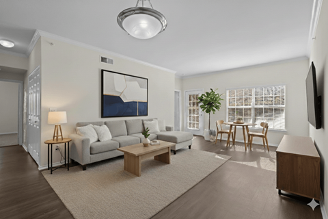 A living room with a grey couch, a coffee table, a lamp, a painting, a dining table with chairs, and a flat-screen TV mounted on the wall.