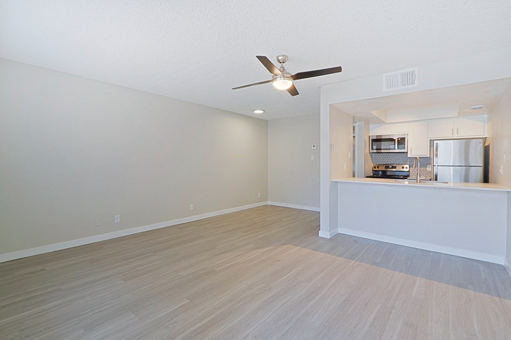 an empty living room with a ceiling fan and a kitchen in the background