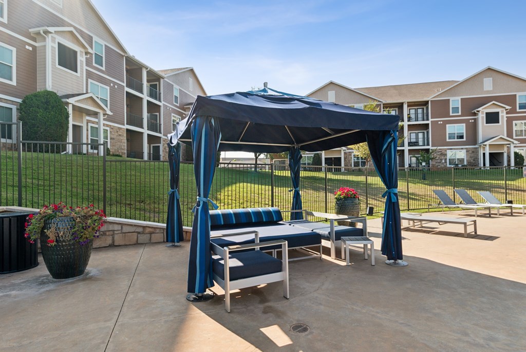 A gazebo with a blue canopy is situated in a courtyard with lounge chairs and potted plants.