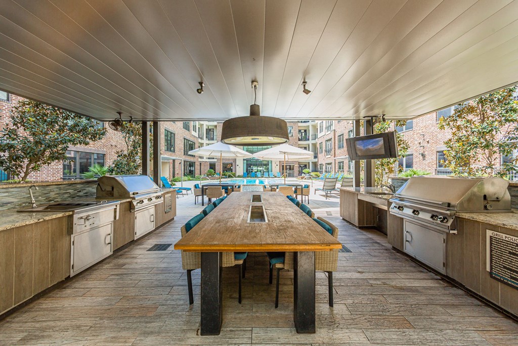 A long wooden table is in the middle of a patio with barbecue grills on either side.