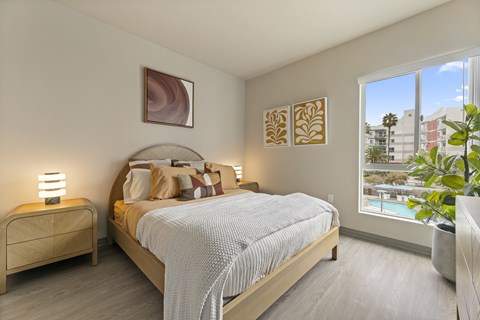 A bedroom with a large bed, a nightstand with a lamp, and a plant.