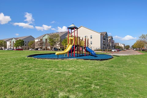 A playground with a slide and a blue mat in the middle of a grassy area.