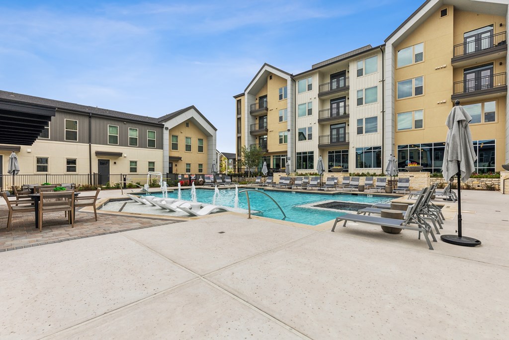 A large swimming pool surrounded by lounge chairs and umbrellas in front of apartment buildings.
