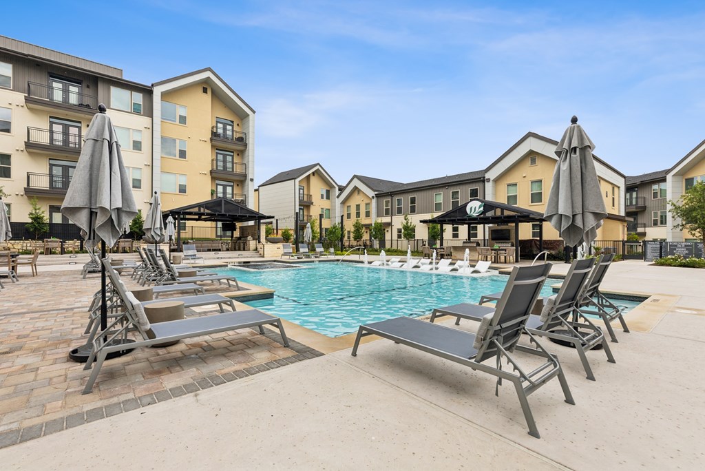 A pool area with sun loungers and umbrellas in front of apartment buildings.