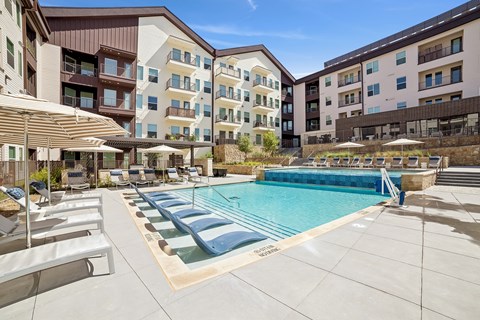 A large swimming pool surrounded by lounge chairs and umbrellas in front of apartment buildings.