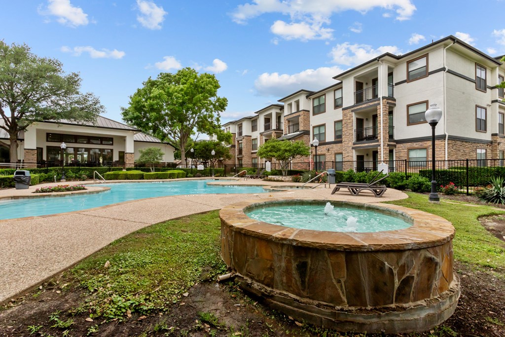 A large outdoor swimming pool with a hot tub in the middle of a grassy area in front of apartment buildings.