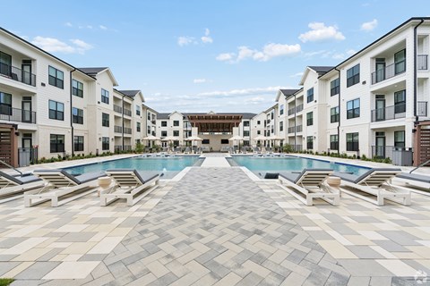 A large swimming pool surrounded by sun loungers in front of apartment buildings.