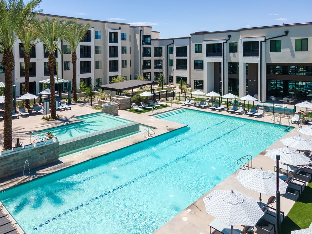 A large swimming pool surrounded by lounge chairs and umbrellas in front of a multi-story building.