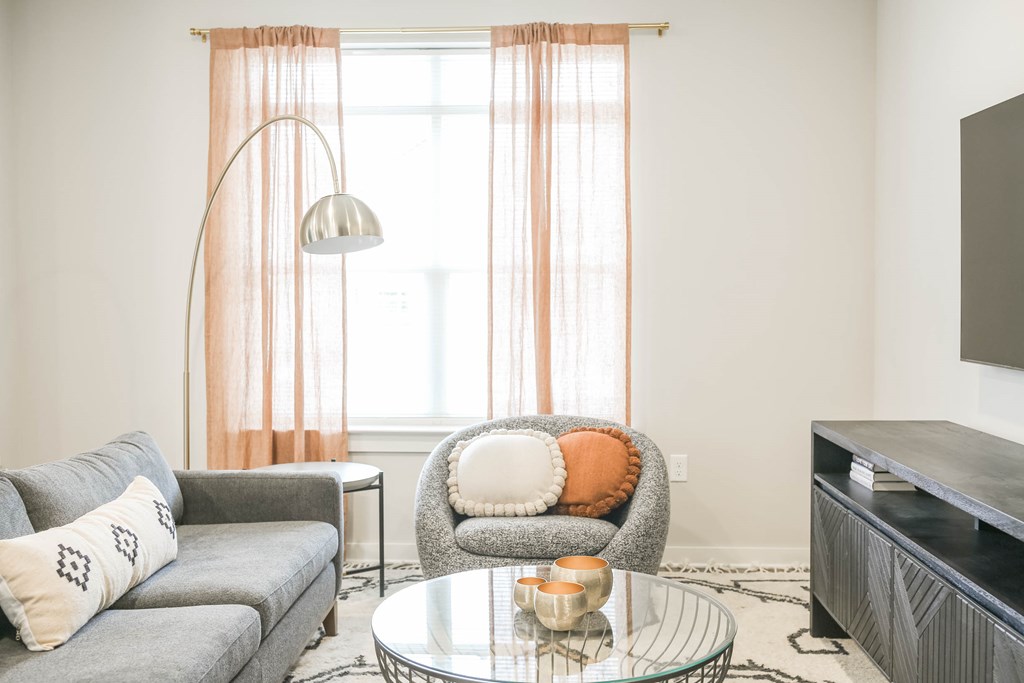 A living room with a grey couch, a glass table, and a grey armchair with orange pillows.