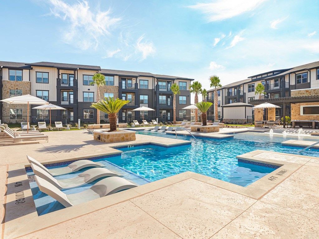 A swimming pool surrounded by lounge chairs and umbrellas in front of a multi-story apartment building.
