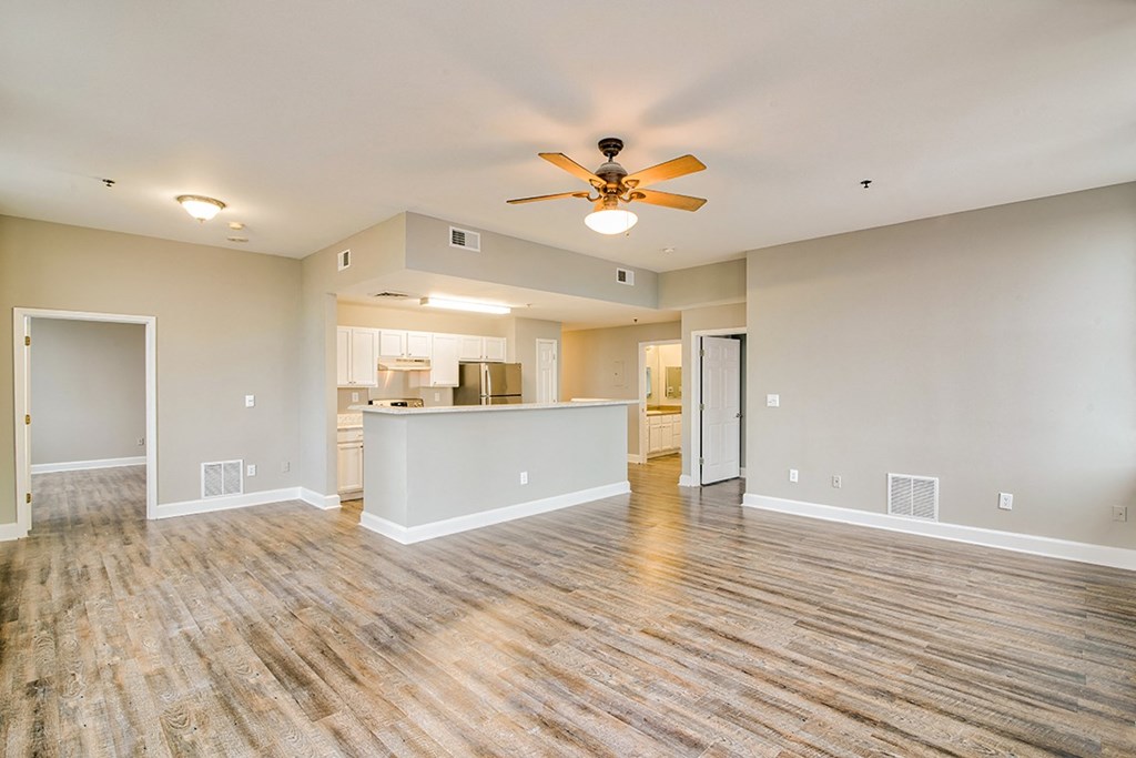 an empty living room with a ceiling fan and a kitchen in the background