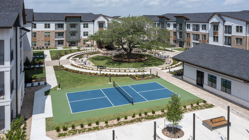 A tennis court is surrounded by a grassy area and apartment buildings.
