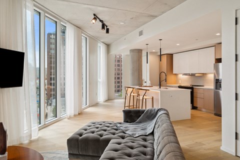 A modern living room with a grey tufted sofa and a kitchen area in the background.