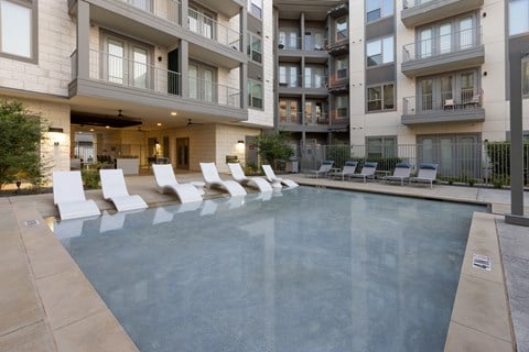 A swimming pool surrounded by white lounge chairs in front of apartment buildings.