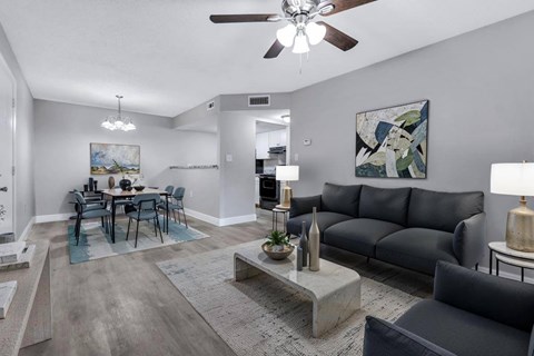 A modern living room with a grey sofa, a grey coffee table, a dining table with chairs, and a ceiling fan.