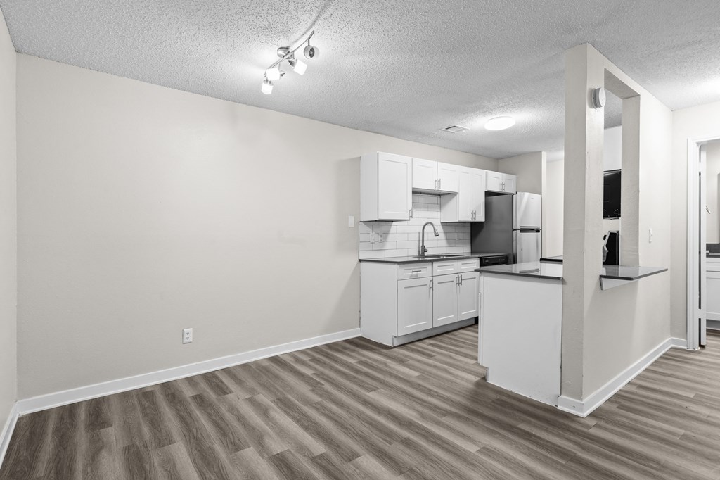 the living room and kitchen of an apartment with a wood floor and white cabinets