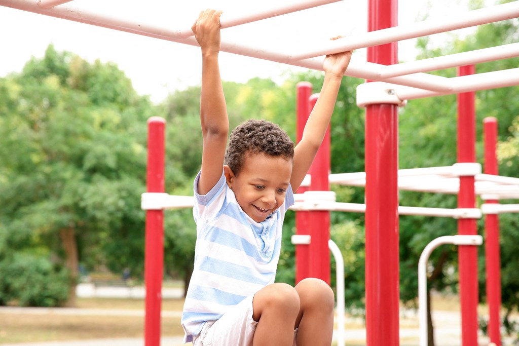 A child in a blue and white striped shirt is hanging from a red and white monkey bars.